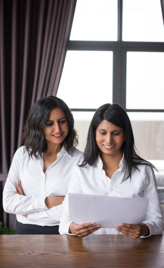 Two smiling female coworkers looking through document in cafe. They are standing with window in background. Document concept. Front view.